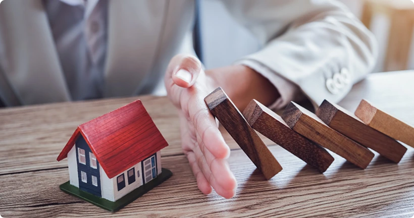 A person in a business suit stops falling wooden blocks from hitting a small toy house.