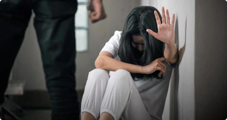 A woman cowering on the floor holds up a hand defensively toward a man with a raised fist.
