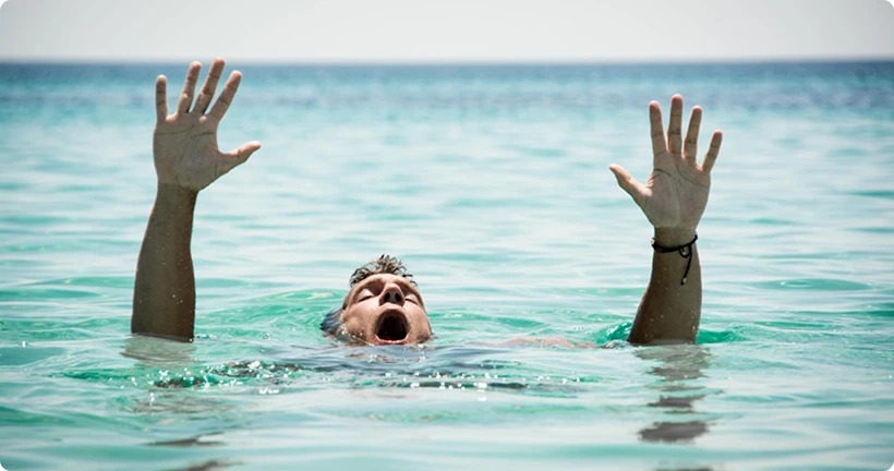 A man in the ocean with his head partially submerged, mouth open, and both hands raised in the air as if calling for help.