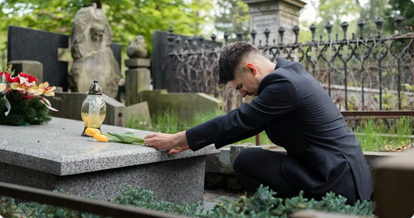 A man in a black suit kneeling by a gravestone to lay down a flower.