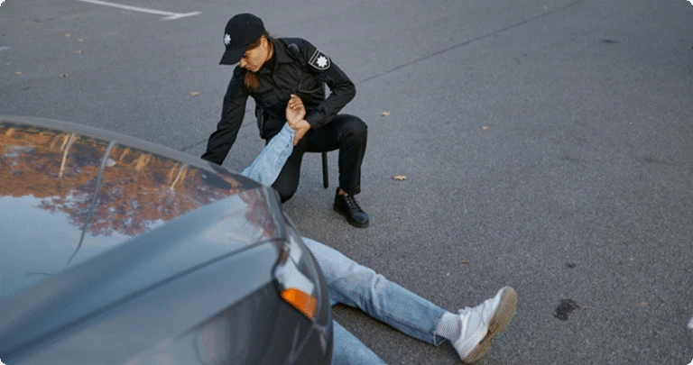 A police officer kneeling next to an injured pedestrian lying on the asphalt in front of a car.