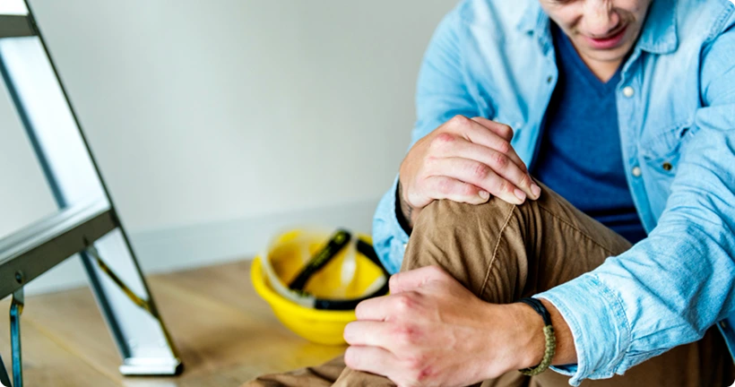 A man sitting on the floor in pain, clutching his injured knee near a step ladder and a yellow hard hat.
