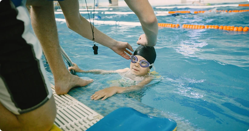An instructor adjusts a young child's goggles and swim cap at the edge of an indoor pool.