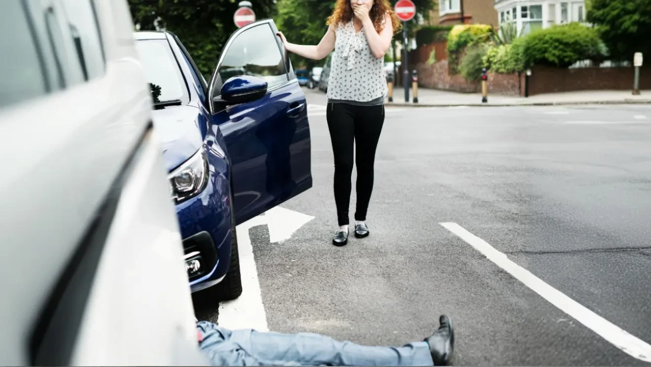 A shocked woman standing by an open car door looking down at an injured pedestrian lying on the street.
