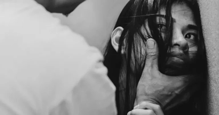 A distressed woman pinned against a wall while a person's hand forcefully covers her mouth.