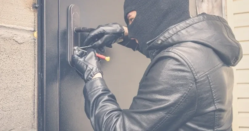 A person wearing a black balaclava and gloves uses lockpicking tools to break into a door.