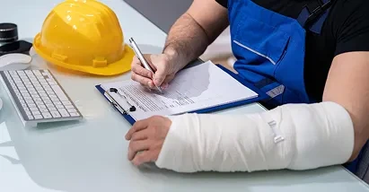 A worker with an arm cast fills out a document on a clipboard next to a yellow hard hat.