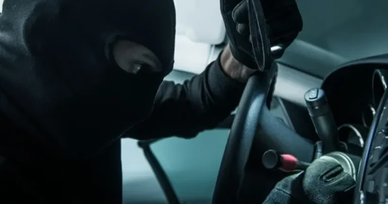 A person wearing a Black balaclava and gloves leans into a car to break into the steering column ignition.